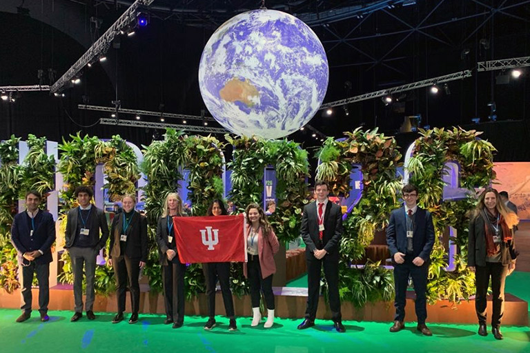 A student and conference attendees stand in front of a sign for the UN Climate Change Conference 2021 while holding an IU flag