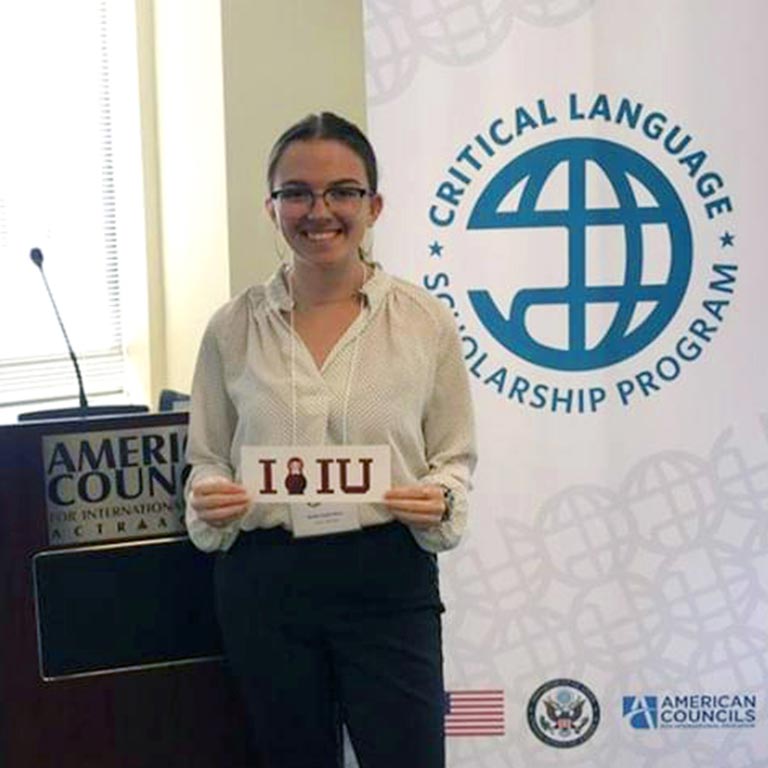 A student holds an IU sign in front of a podium with a banner for the Critical Language Scholarship Program in the background