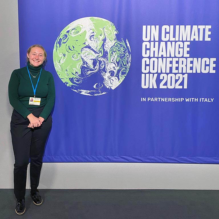 A student wearing a credential badge stands in front of a sign for the UN Climate Change Conference 2021
