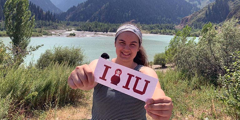 Student holds up an IU sign while posing in front of a lake and mountain vista on a study abroad trip